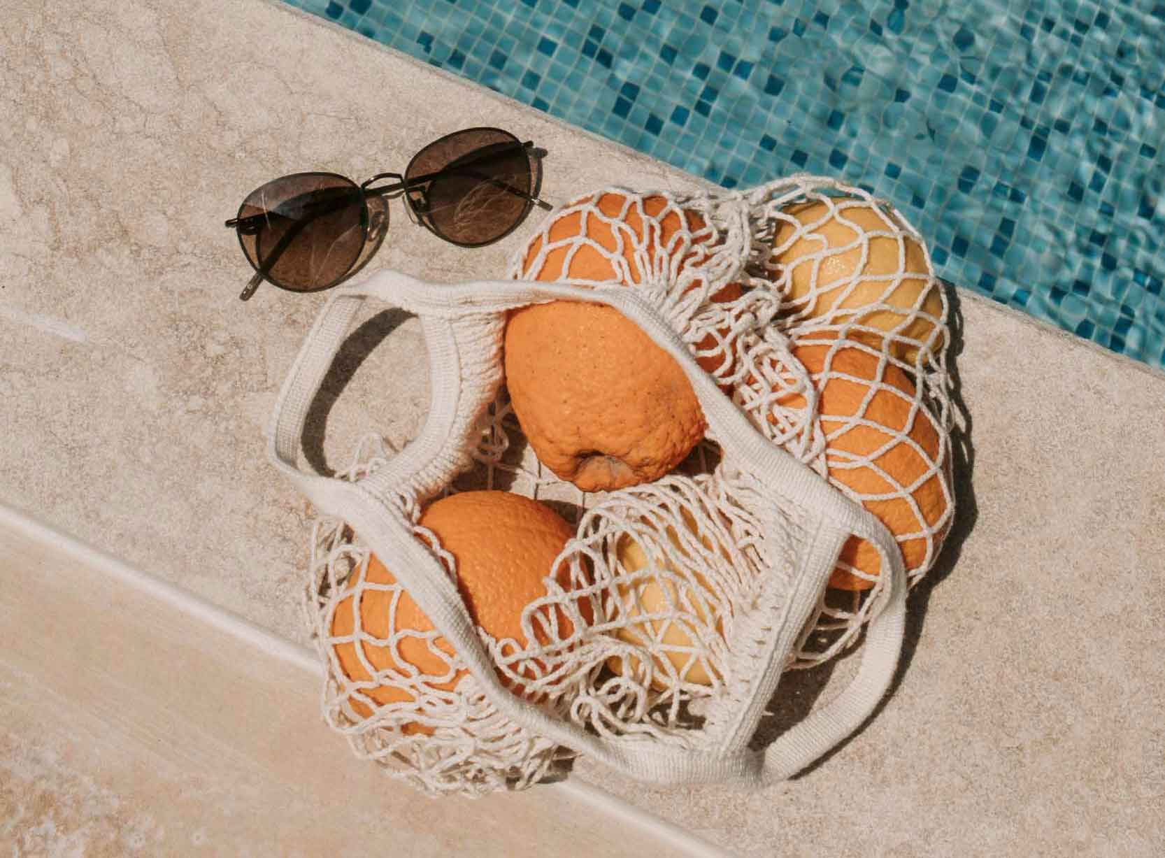 A basket of oranges and sunglasses rests on a ledge beside a sparkling swimming pool, evoking a summer vibe