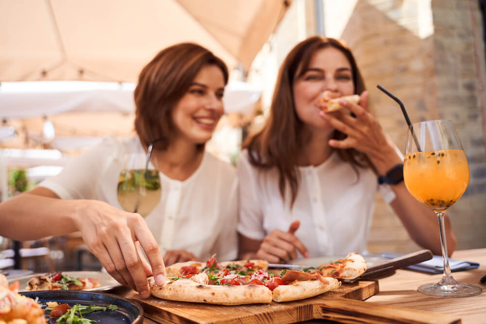 Two women enjoy pizza and drinks at an outdoor restaurant patio.