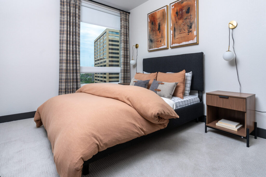 Two Bedroom Luxury Apartments in Denver, CO 1 Modern bedroom with a large window, plaid curtains, a dark upholstered bed with tan bedding, abstract wall art, and a wooden nightstand.