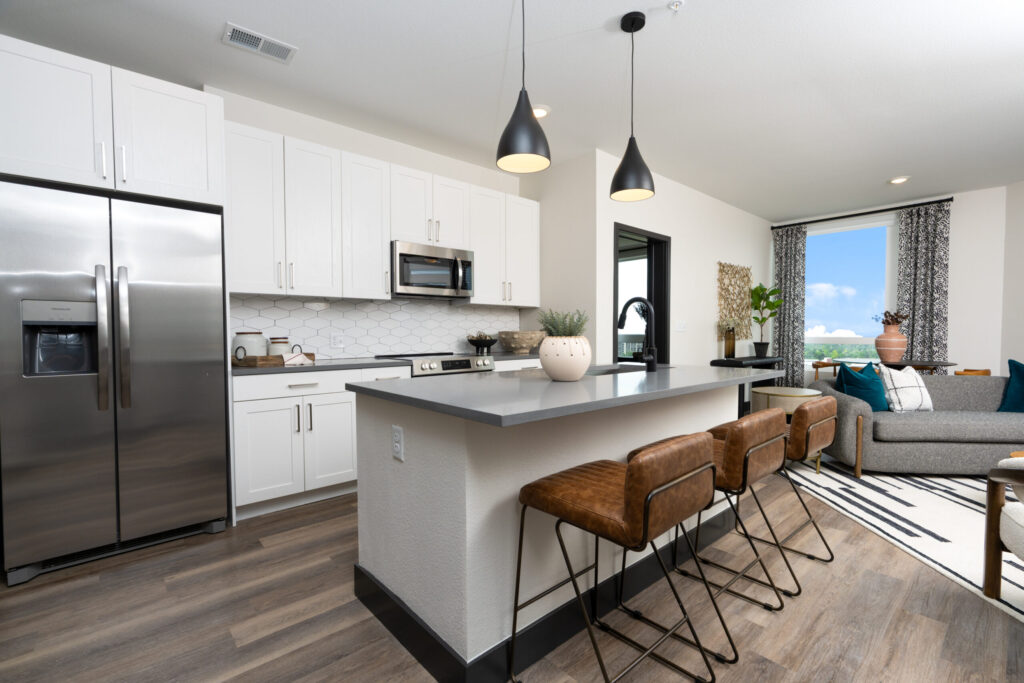 Two Bedroom Luxury Apartments in Denver, CO 2 Modern apartment kitchen with white cabinets, stainless steel appliances, a gray countertop island with brown barstools, and a bright living area with large windows.