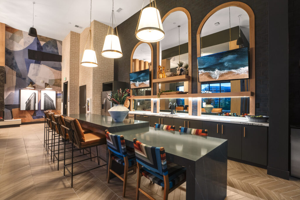 Two Bedroom Luxury Apartments in Denver, CO 3 Stylish community lounge with a long island counter, modern pendant lighting, colorful chairs, large mirrors, and mounted TVs above the bar area.