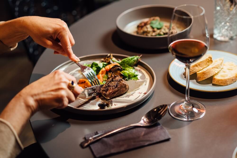 Person enjoying a plated steak dinner with vegetables and a glass of red wine at a restaurant.